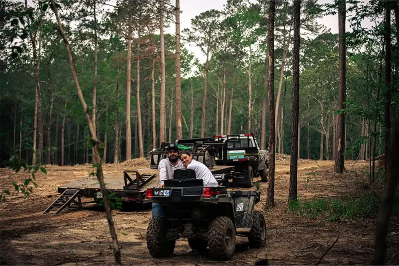 A couple sitting together on a utility vehicle in a clearing among tall pines, with a truck and trailer behind them