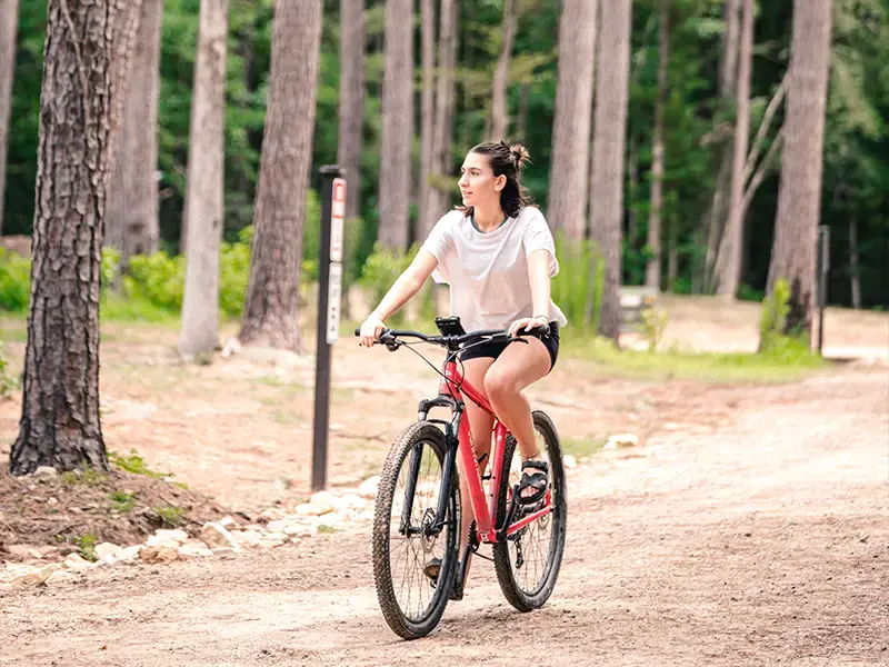 A young woman rides a red bike down a sandy path through the pines