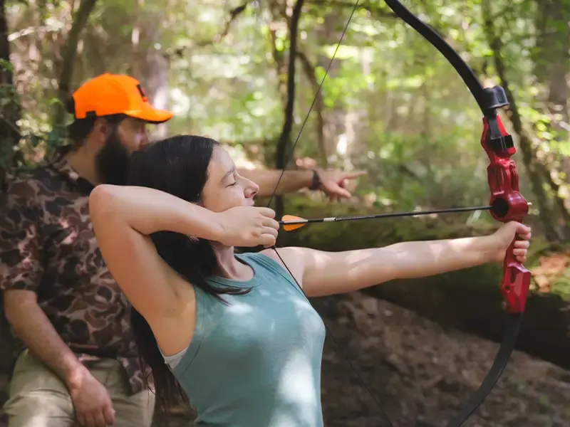 A woman draws a red bow with an arrow nocked while a man in an orange cap points out the target
