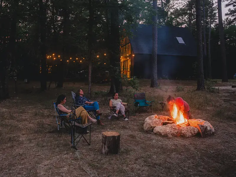 Friends in camp chairs around a stone fire ring at dusk, string lights glowing on a cabin behind them