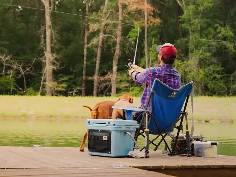 A man in a plaid shirt and red cap casts a line from the dock, his dog beside a cooler and camp chair