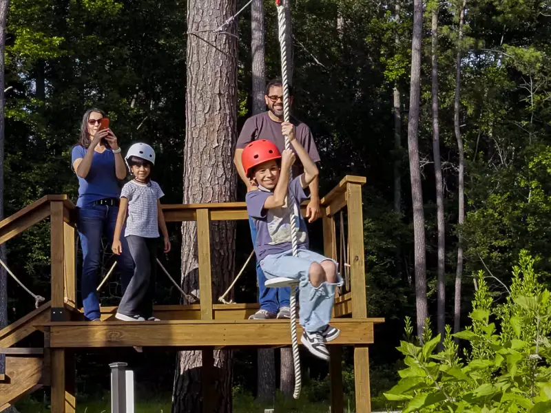 A boy in a red helmet sits on the glider rope seat at a wooden launch platform as his family watches