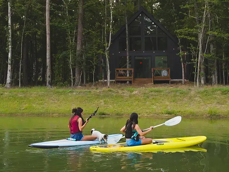 Two girls paddle kayaks across the pond, a small dog riding along, with a black A-frame cabin in the trees behind them