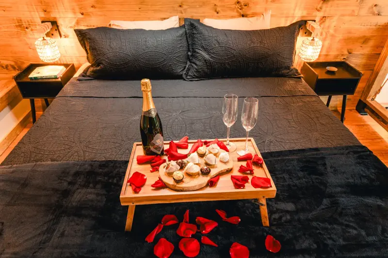A cabin bed with a wooden tray of chocolates, a bottle of sparkling wine, two glasses and red rose petals