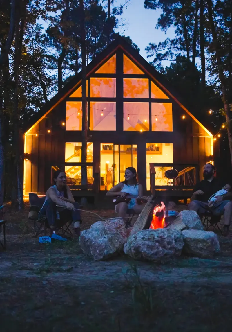 A lit A-frame cabin at dusk with three guests around a campfire out front, one playing guitar