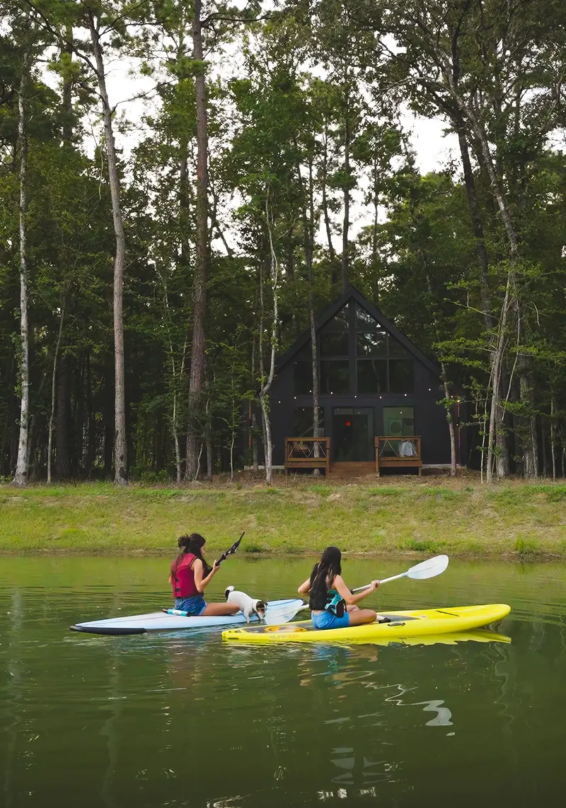 Two girls on a kayak and a paddleboard with a small dog on the pond, a cabin in the pines behind them