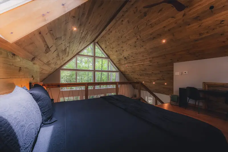 Loft bedroom under a vaulted wood ceiling with a triangle window looking into the trees