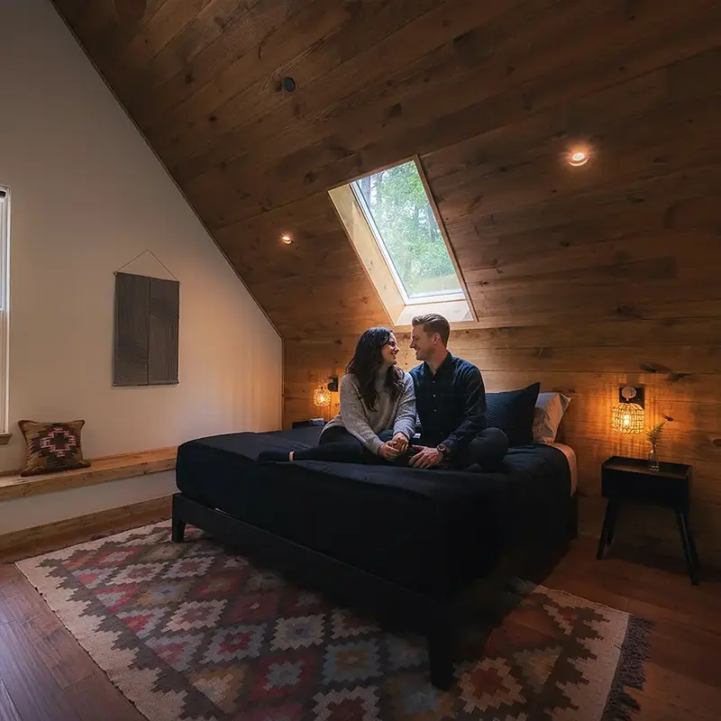A couple sitting together on a dark-quilted bed under a skylight in a wood-paneled cabin loft