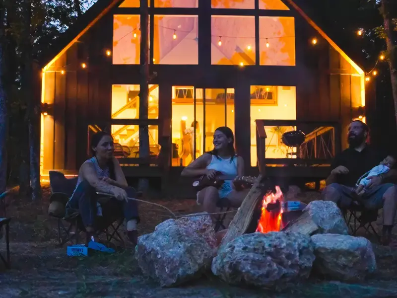 Family around a stone fire pit at dusk, one playing guitar, a cabin with glowing windows behind them